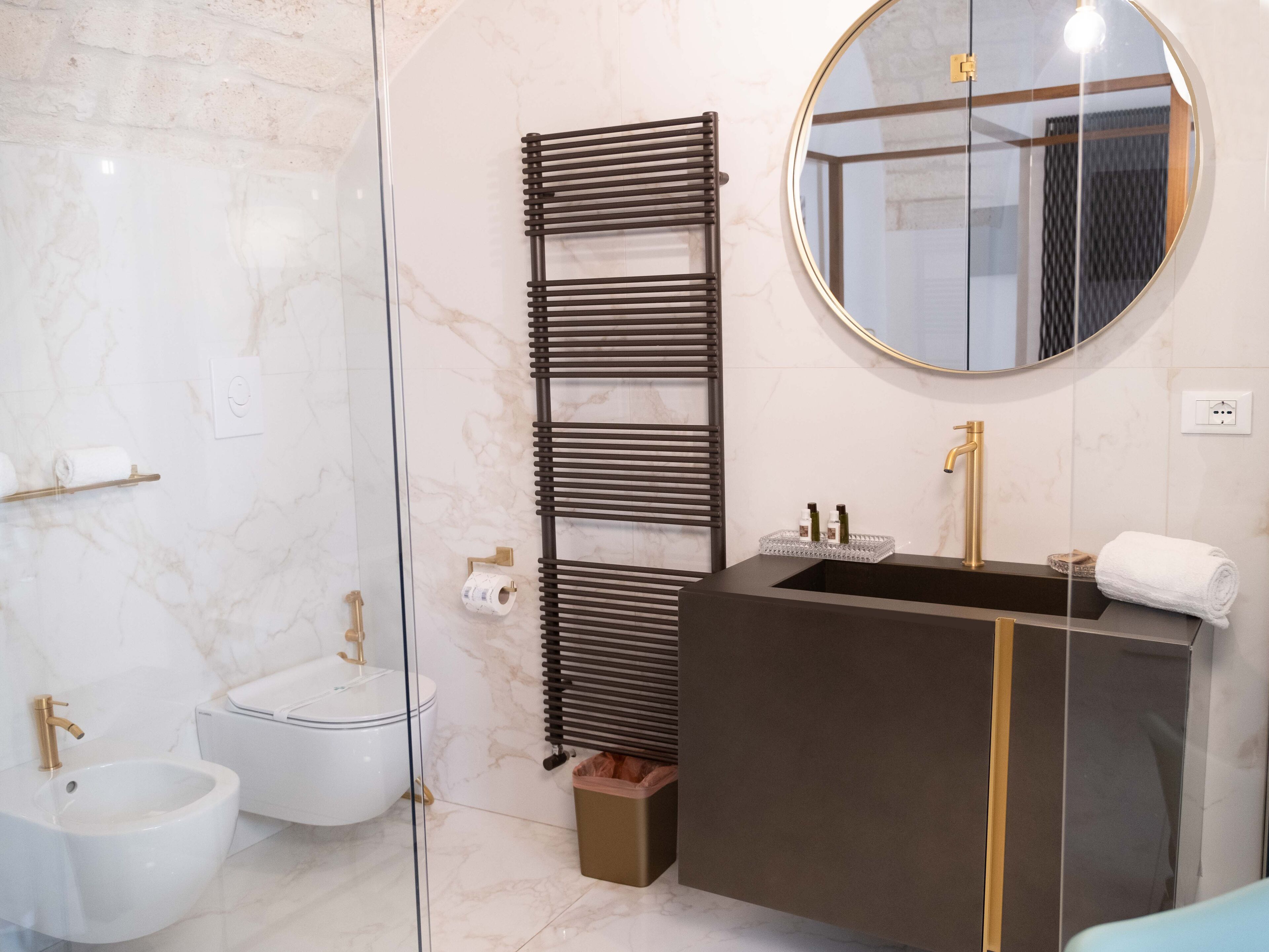 A modern bathroom with a glass shower enclosure, a black towel rack, a round mirror, a gold faucet sink, and a bidet.