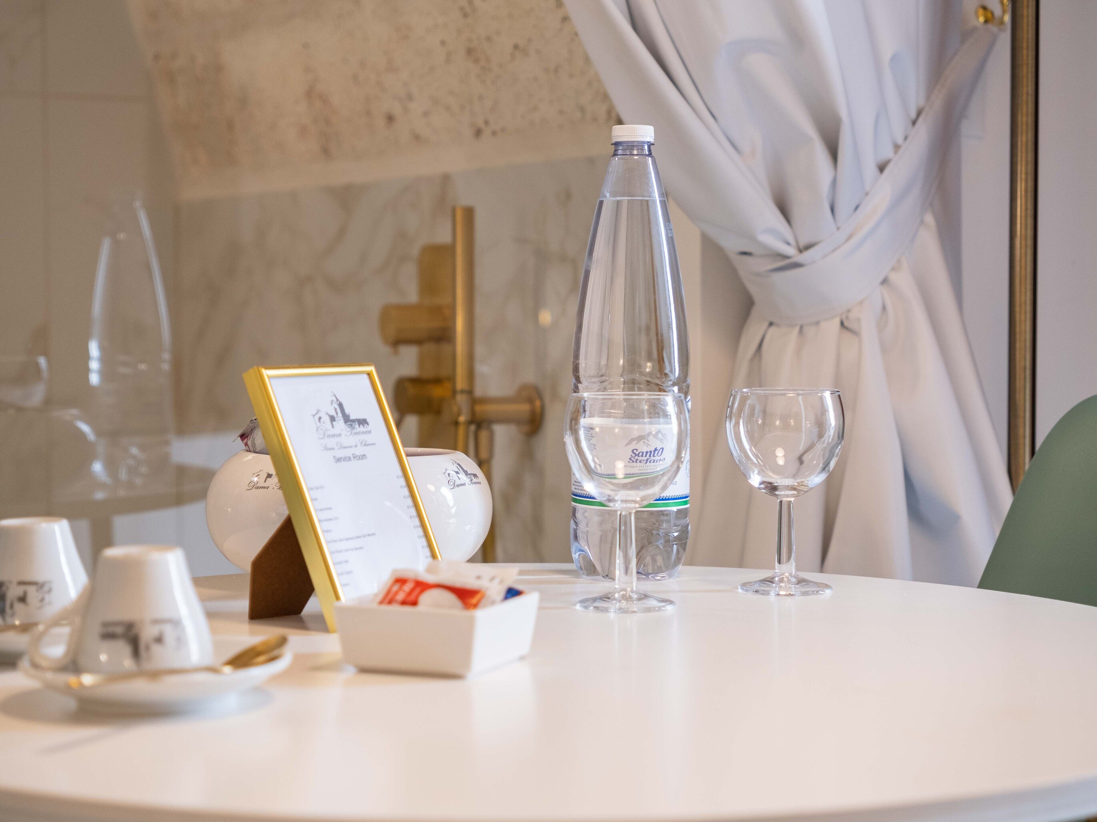 A hotel room with a round table set for two, featuring a bottle of water, two wine glasses, a menu, and a small box of tissues.
