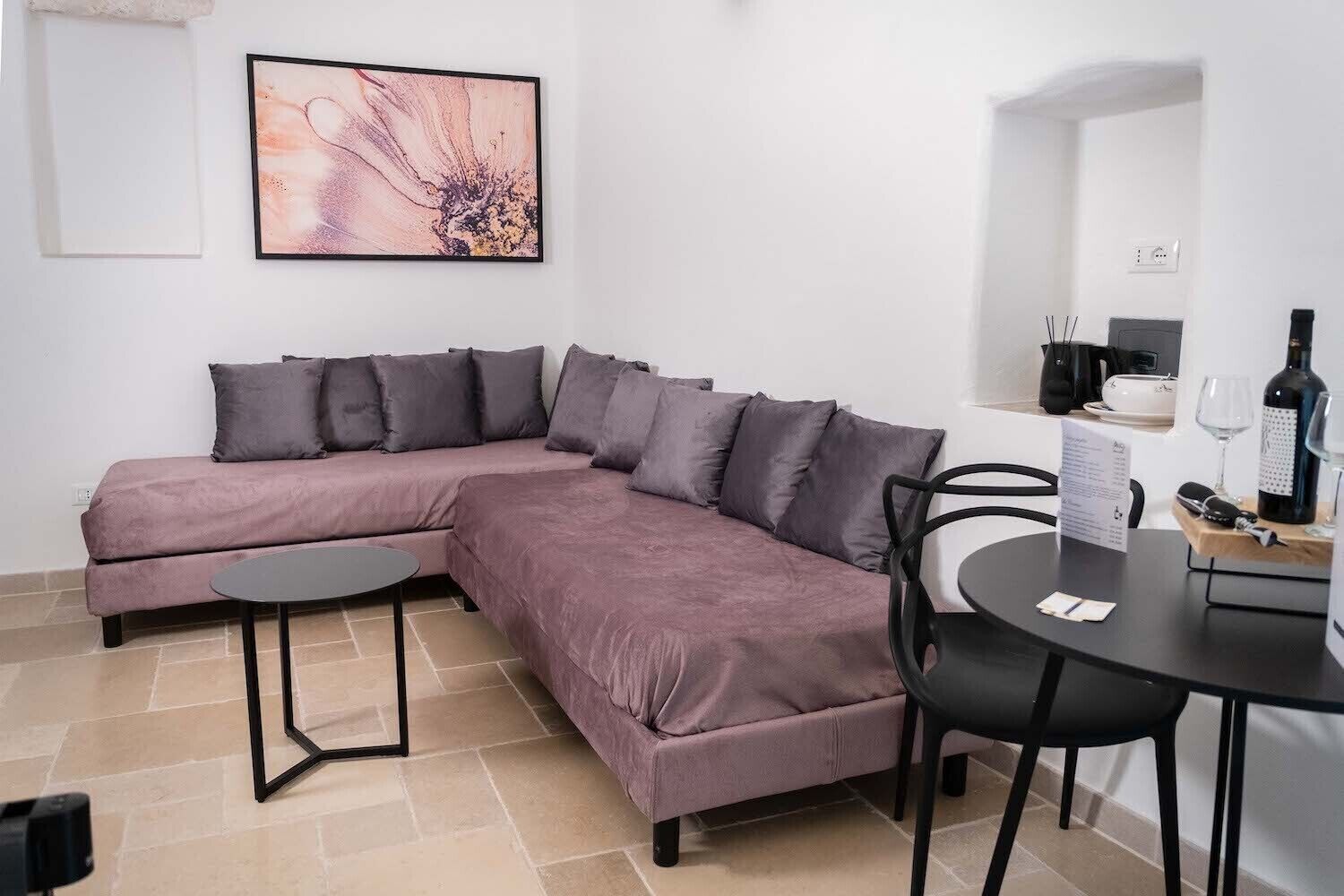 Ein modernes Wohnzimmer mit einer L-förmigen Couch, einem kleinen runden Tisch und einem Essbereich mit einem Stuhl und einer Weinflasche.