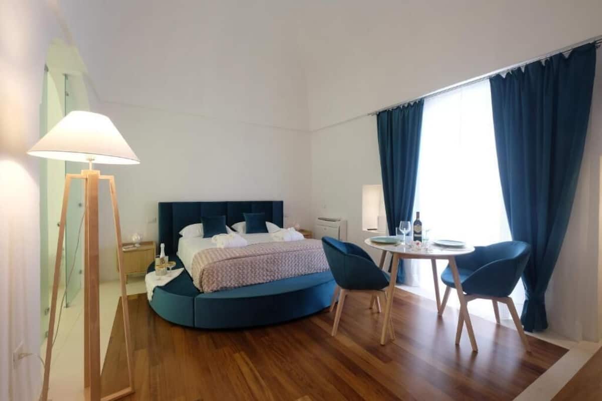 Habitació d'hotel moderna amb un llit rodó, una taula petita amb cadires, una làmpada i vista a l'exterior a través de grans finestres.