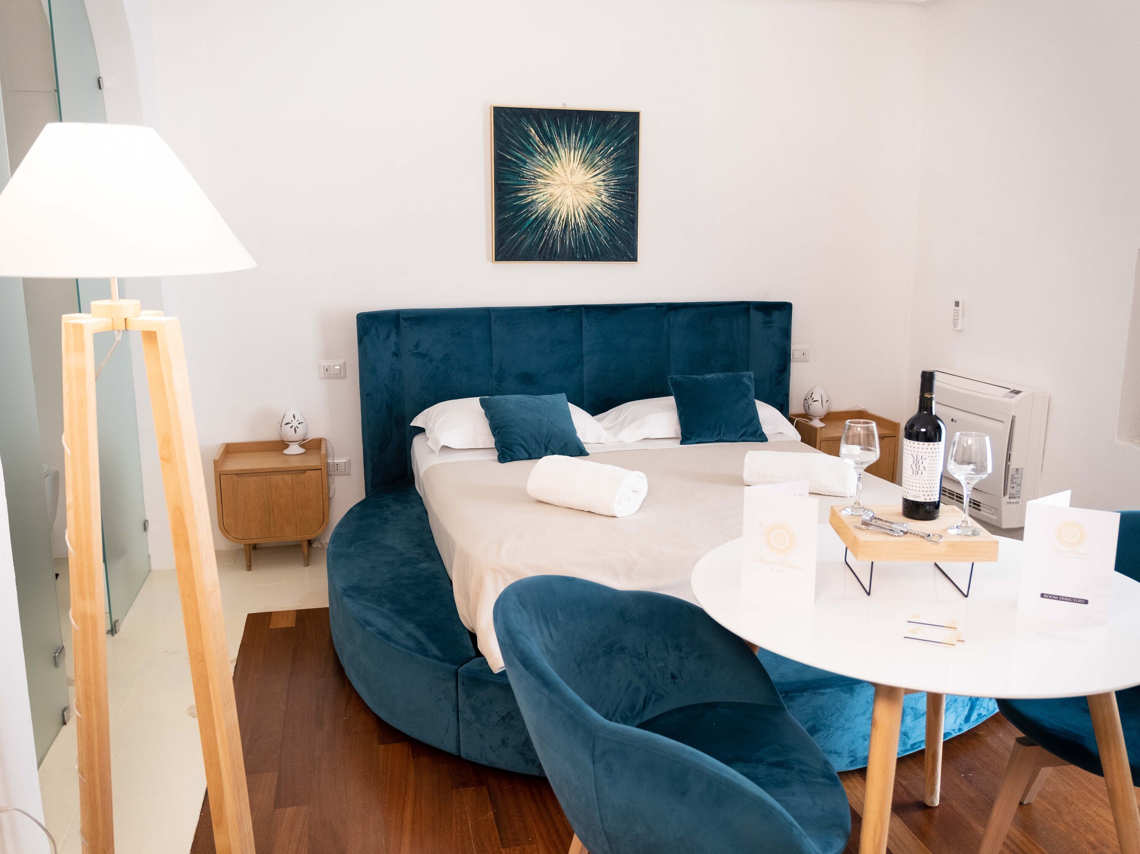 A modern bedroom with a large bed, a bedside table, a lamp, a painting, a wine bottle, and a glass on a small table.