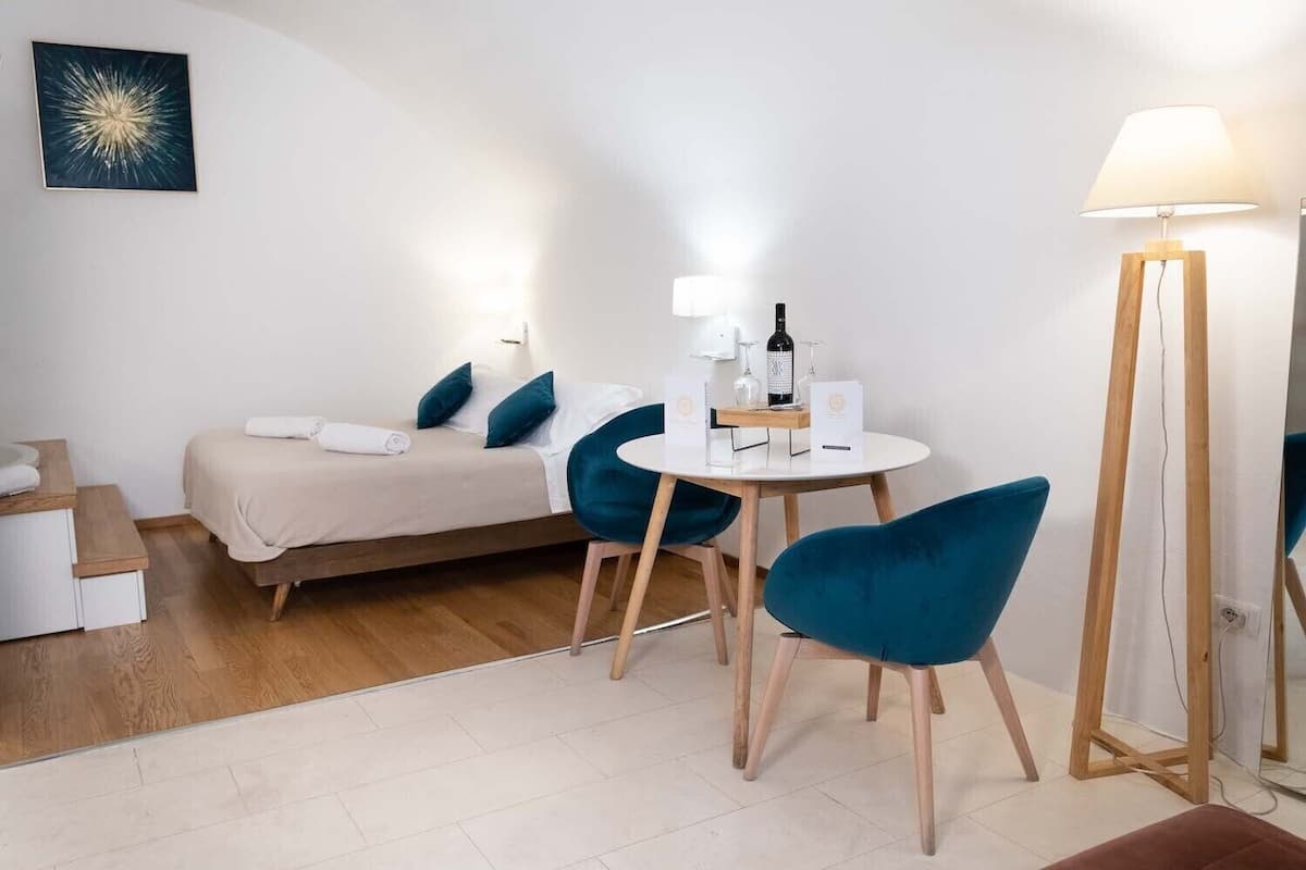 Habitació petita i moderna amb llit, taula rodona i cadires, làmpada de peu i una obra d'art penjada a la paret.