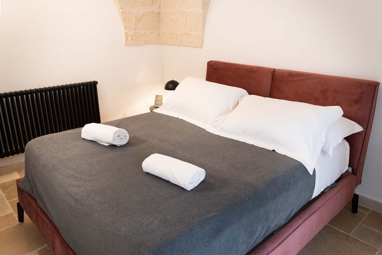 A neatly made bed with a grey bedspread and two rolled white towels, a brown headboard, and a black radiator in the background.