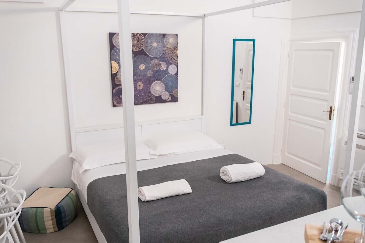 Habitació d'hotel moderna amb llit amb cortines, un quadre a la paret, un mirall i una taula petita amb una ampolla de vi.