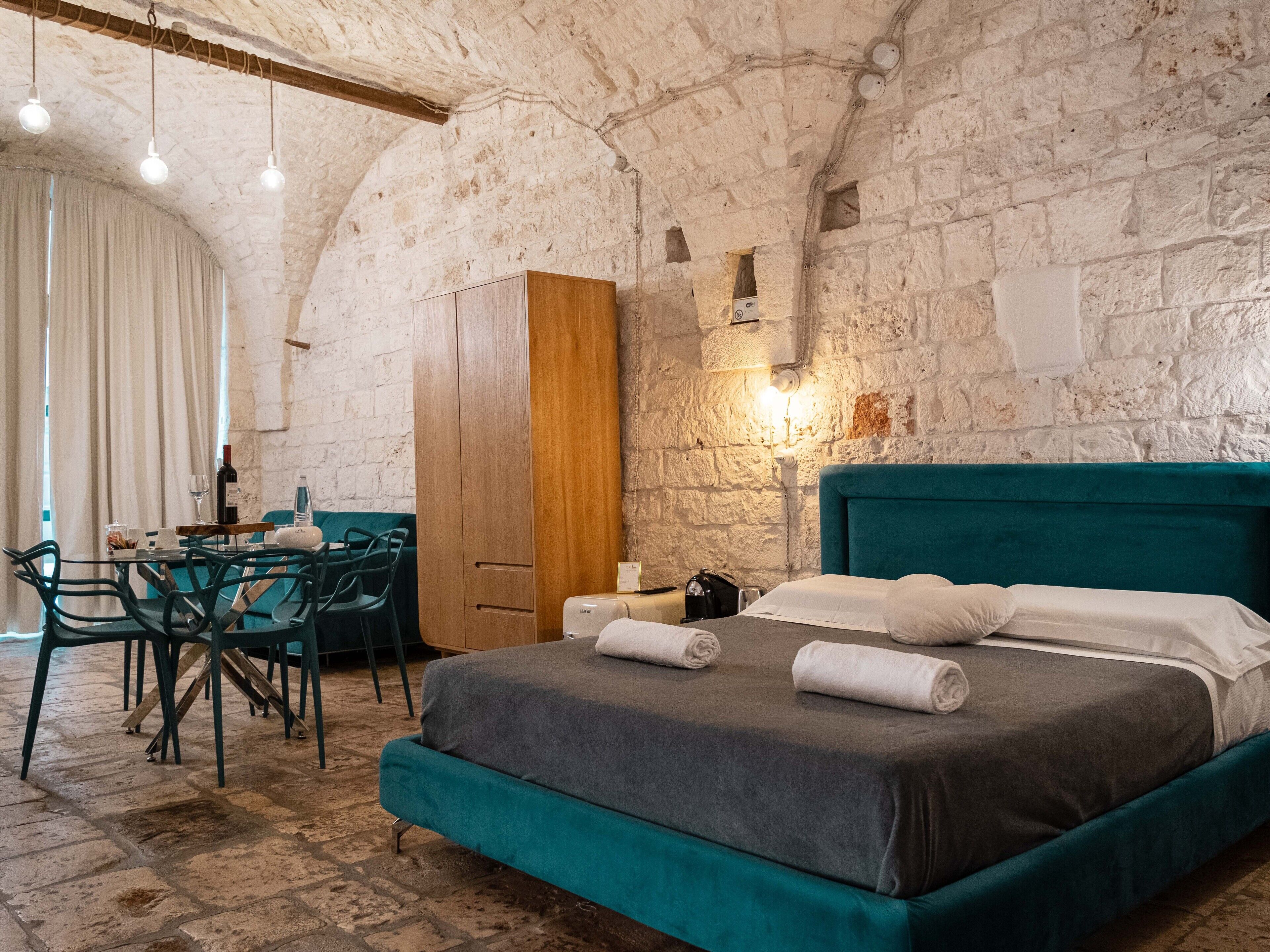 Een kamer met een stenen muur, een bed met blauwgroene beddengoed, een houten garderobe en een tafel met stoelen.