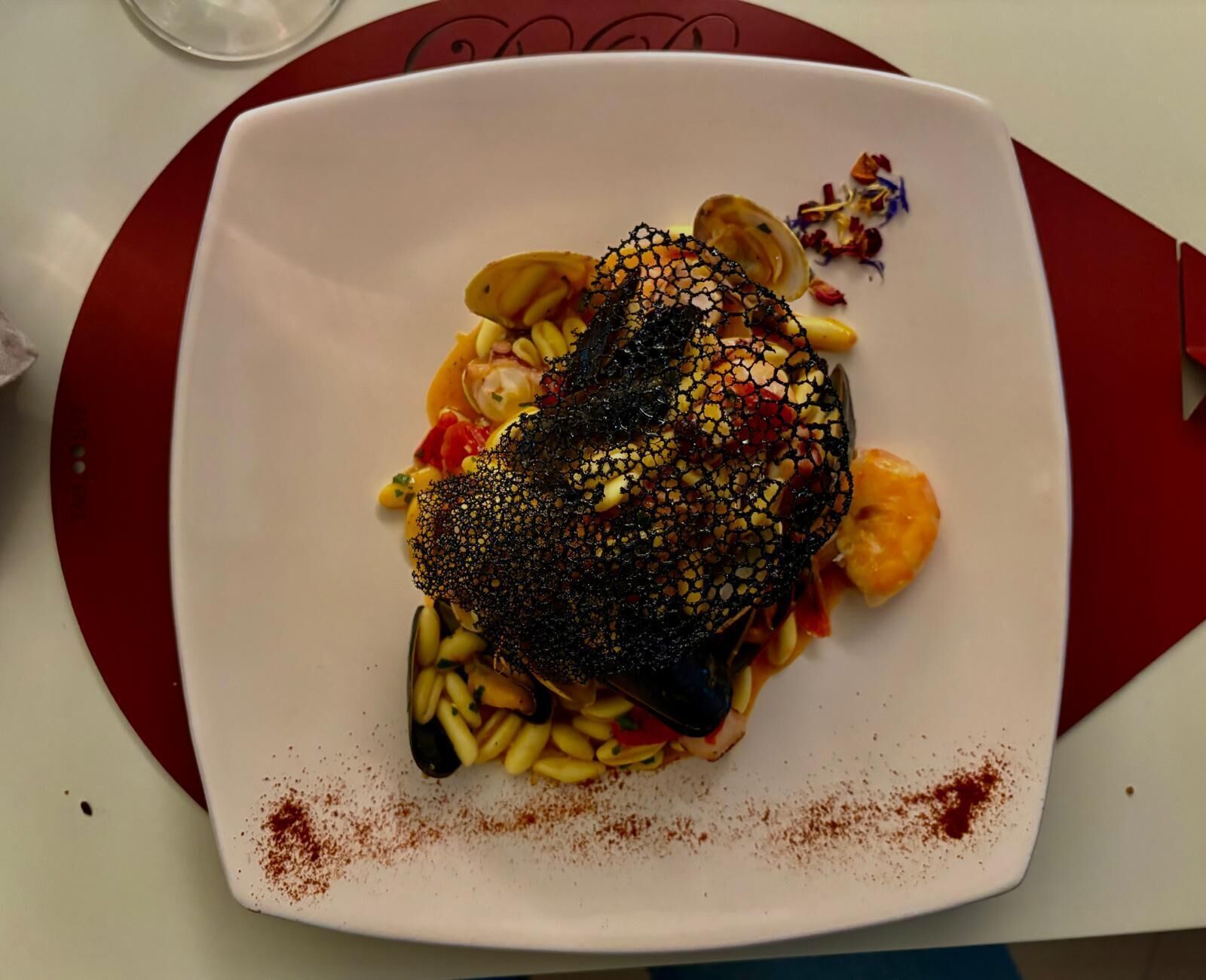 A square plate with a dish of food, including a piece of meat covered in a black net, surrounded by vegetables and garnished with red spice.