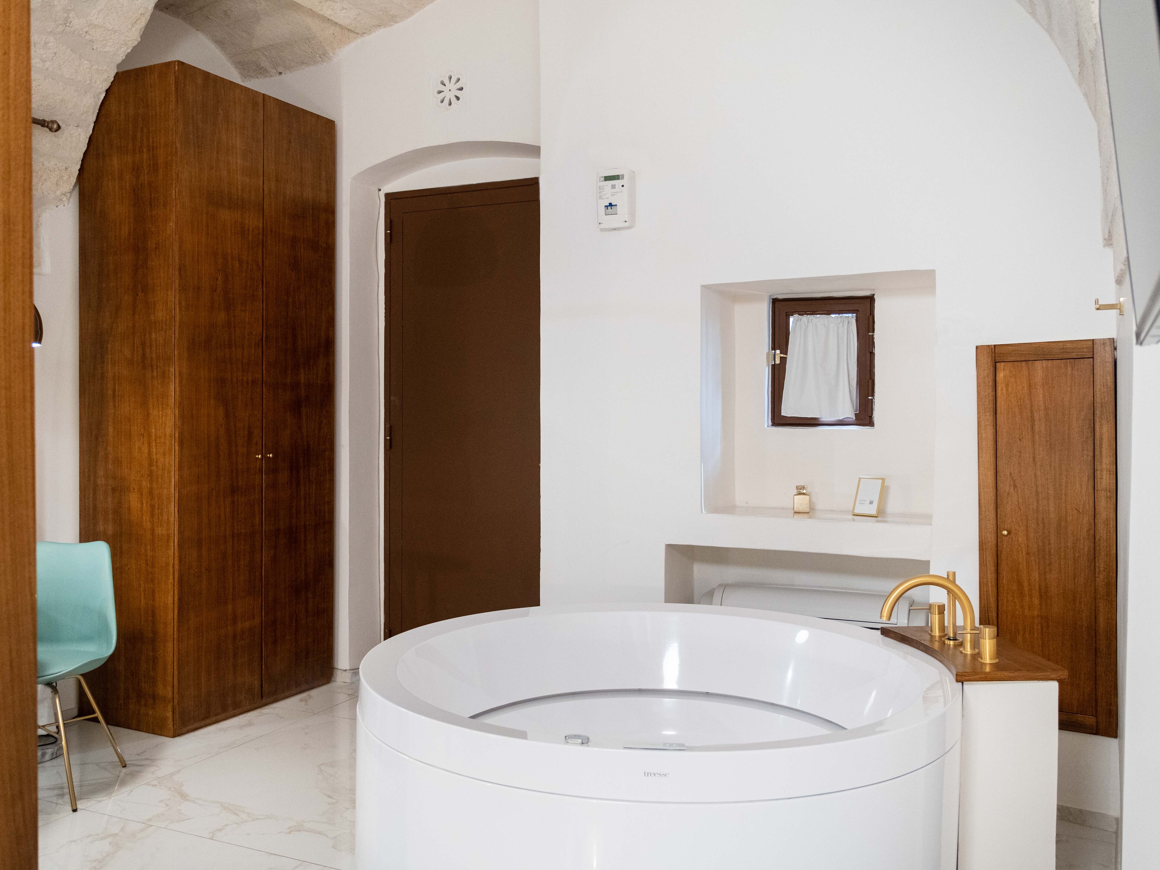 Un baño moderno con una bañera grande, puertas de madera, un ventanal pequeño con una toalla y un lavamanos blanco con detalles dorados.
