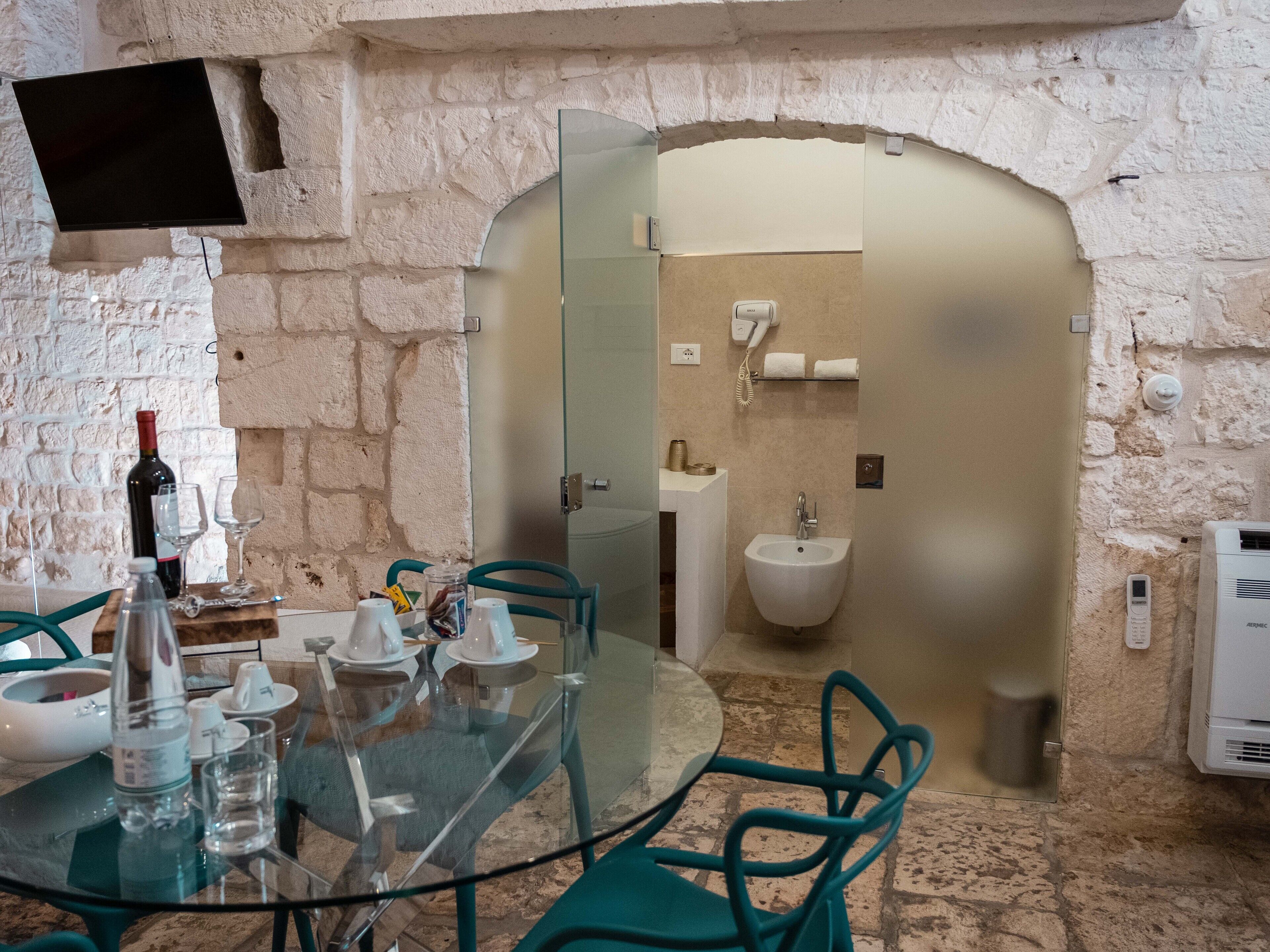 A stone-walled room with a glass table and chairs, a wine bottle, glasses, and a hairdryer on a shelf.