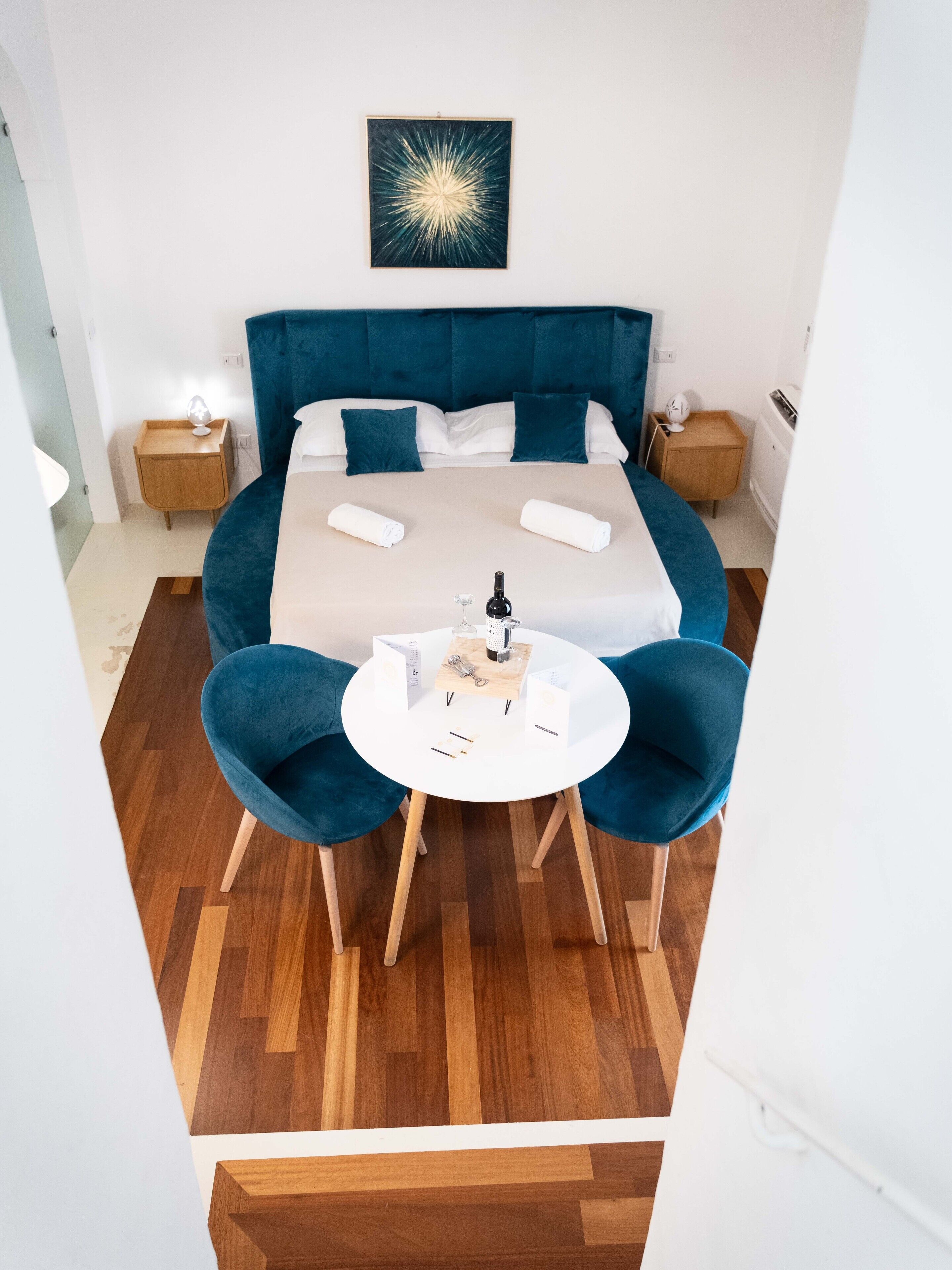 Una habitació d'hotel moderna amb un llit gran, dues taules de nit, una taula rodona amb cadires i una obra d'art penjada a la paret.