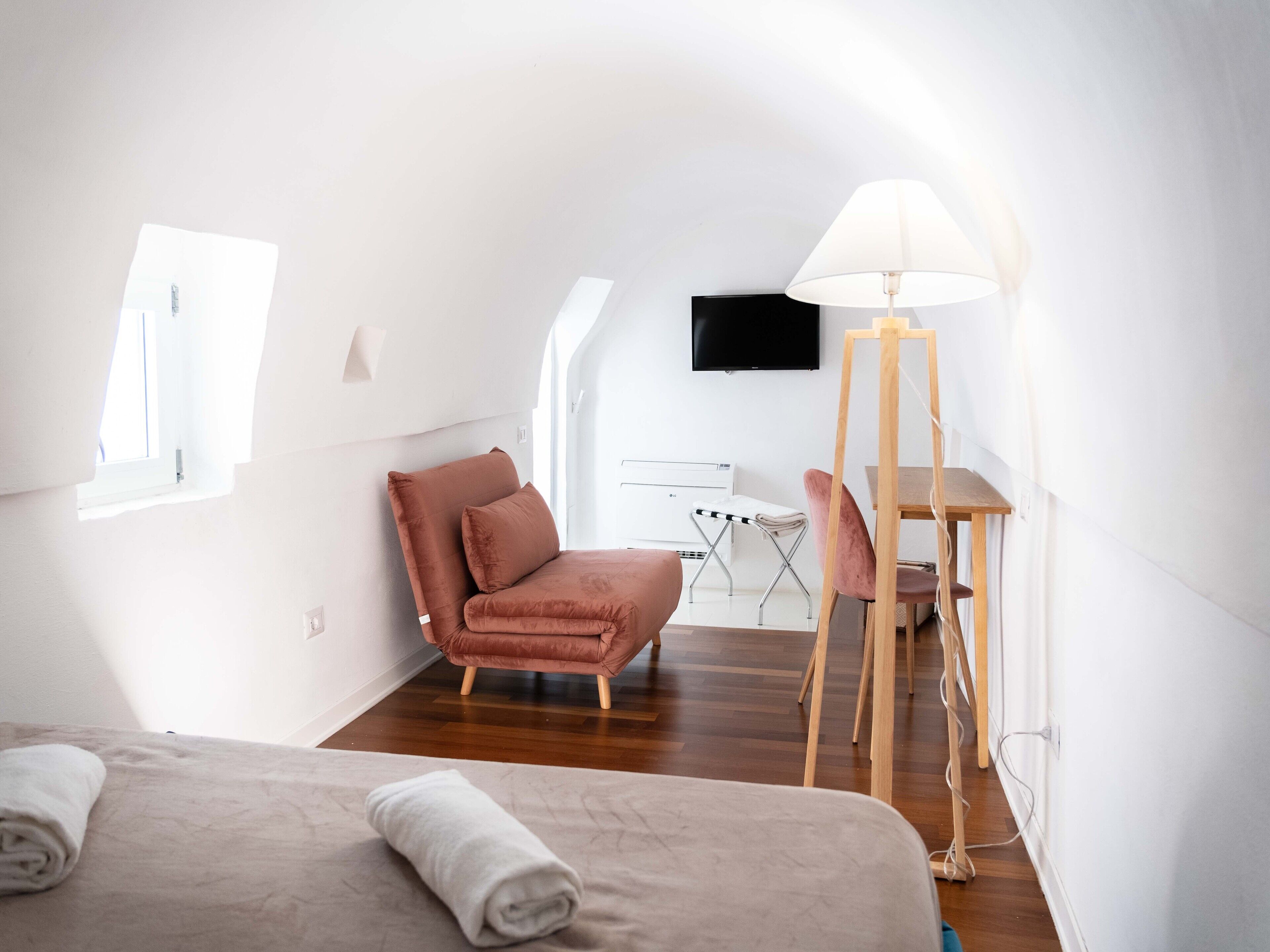 A modern, well-lit room with a bed, a pink chair, a desk with a lamp, and a TV on the wall.