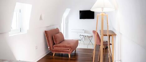 A modern, well-lit room with a bed, a pink chair, a desk with a lamp, and a TV on the wall.