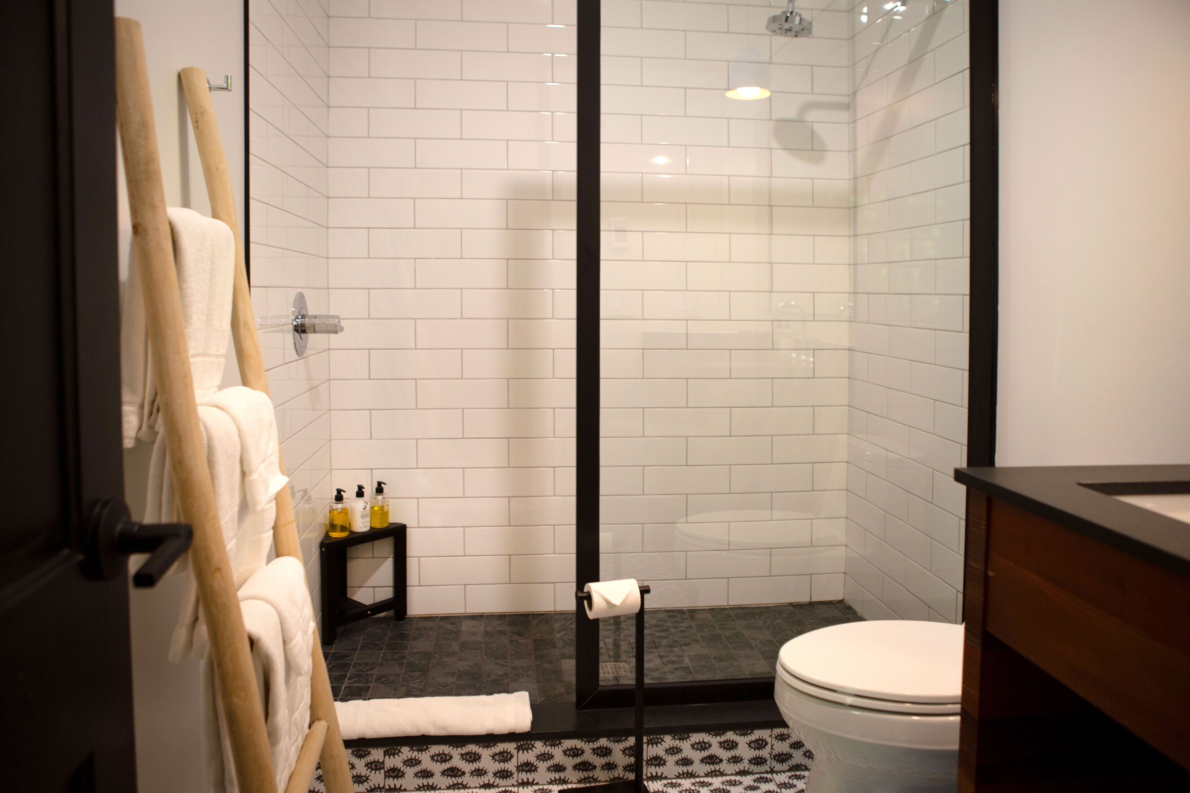A modern bathroom with white tiled walls, a glass shower enclosure, a wooden towel rack, a small table with toiletries, and a white toilet.