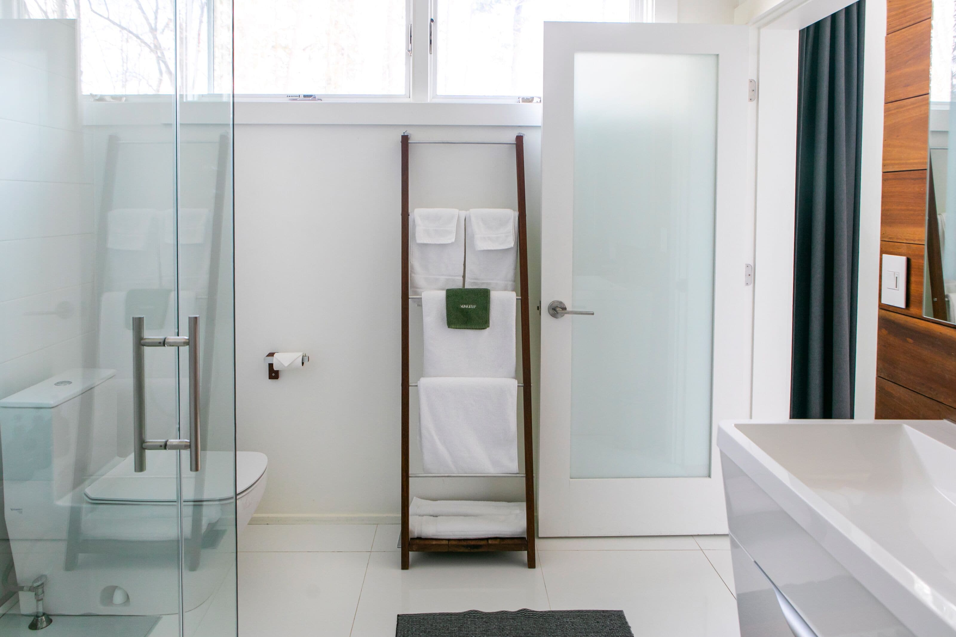 A modern bathroom with a glass shower enclosure, a wooden towel rack with white towels, a white toilet, and a large mirror.