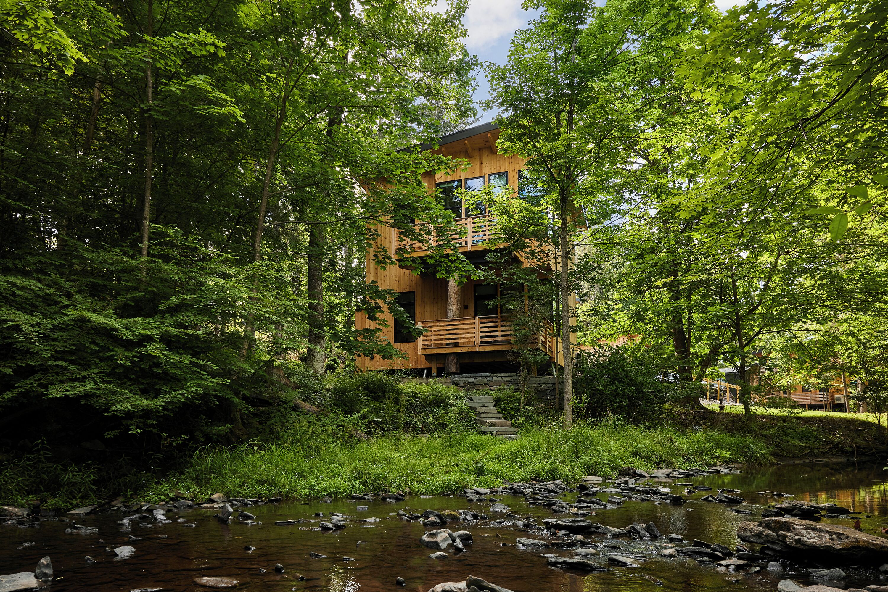 A wooden cabin with a balcony overlooking a forested area and a stream.
