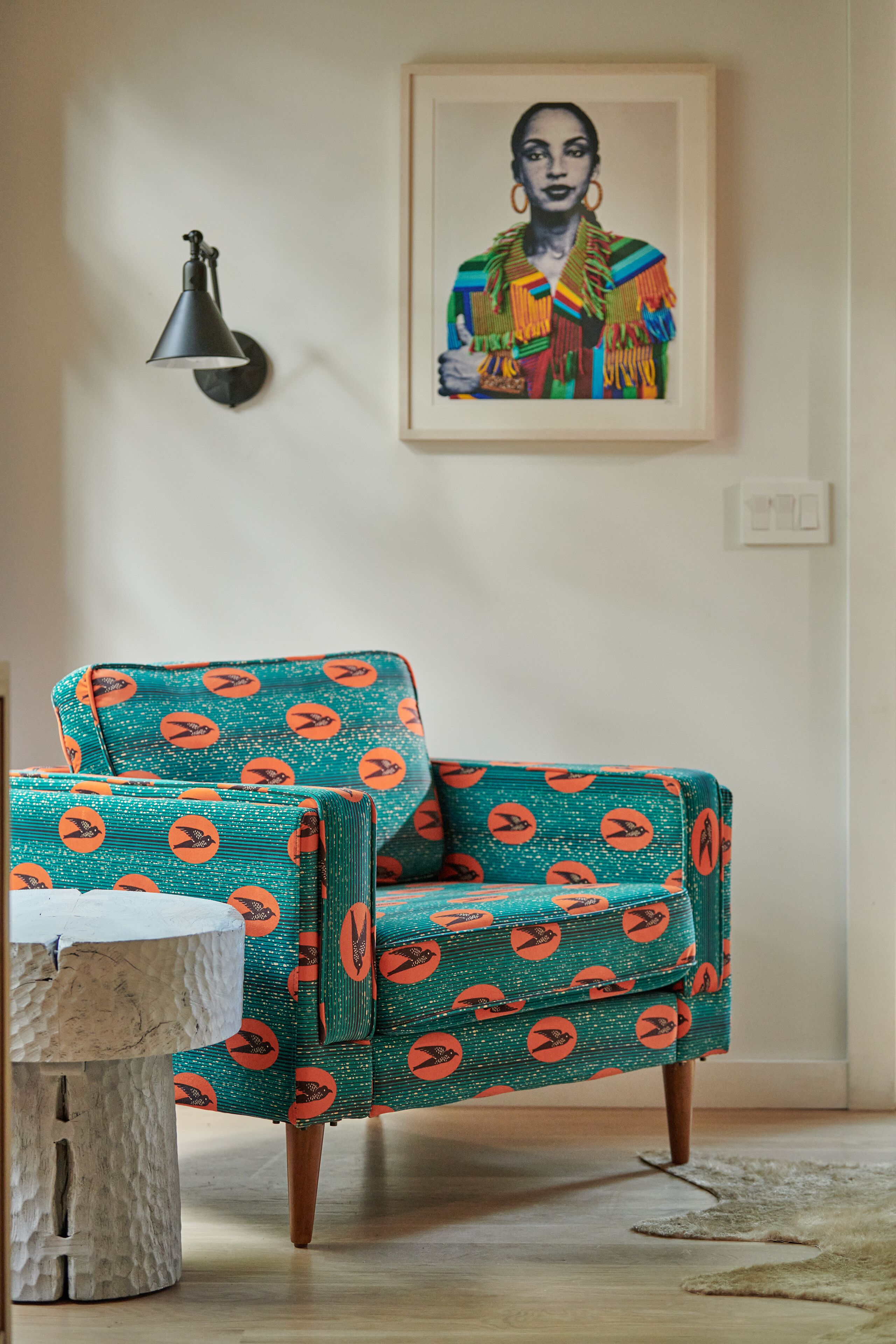 Ein türkis-farbener Sessel mit orangefarbenen Mustern, ein weißer Tisch und eine schwarze Wandleuchte in einem Raum mit einem gerahmten Kunstwerk über dem Sessel.