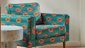 Ein türkis-farbener Sessel mit orangefarbenen Mustern, ein weißer Tisch und eine schwarze Wandleuchte in einem Raum mit einem gerahmten Kunstwerk über dem Sessel.
