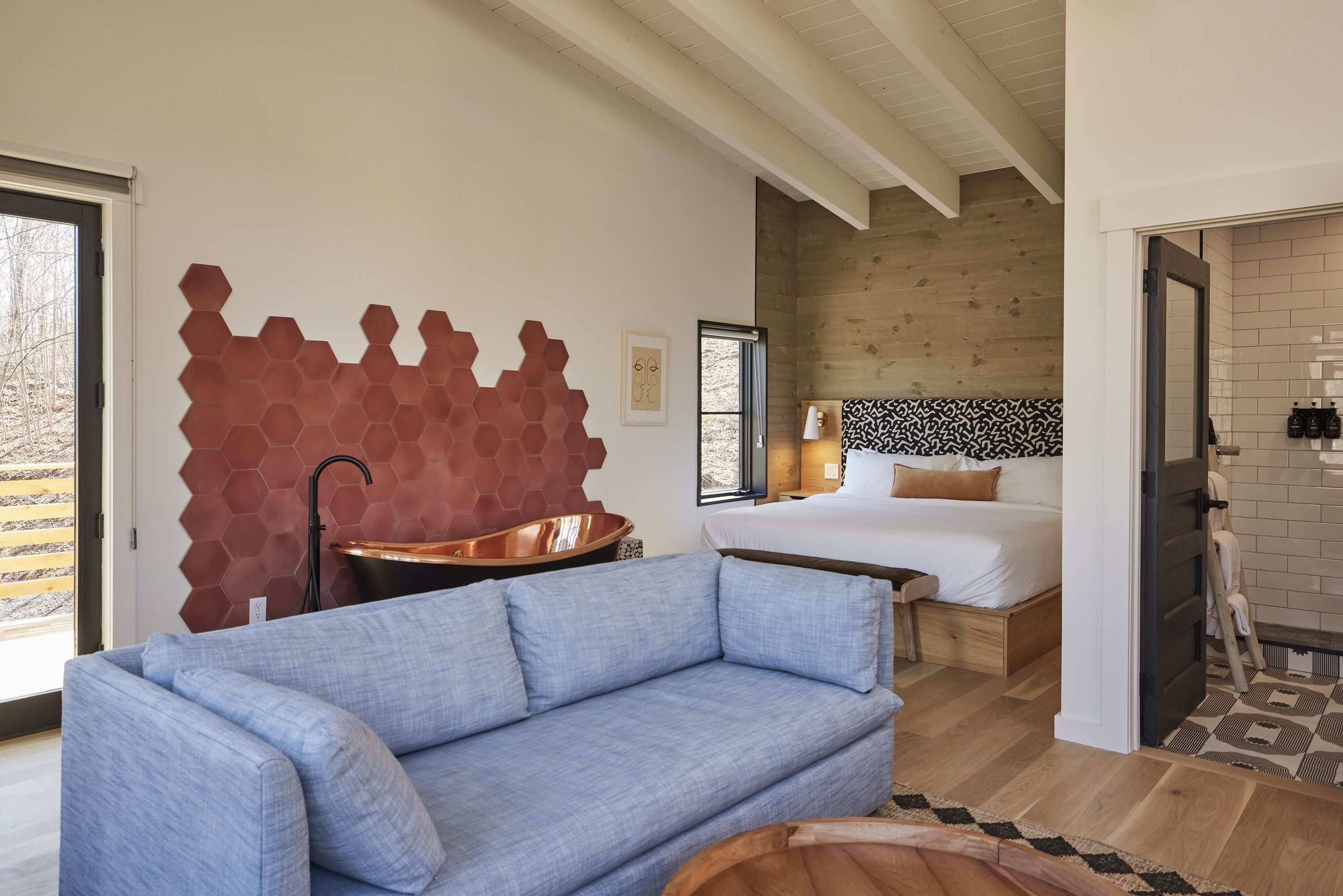 Ett modernt hotellrum med en stor säng, en blå soffa, ett handfat i koppar och en unik väggdesign i form av en hexagon.