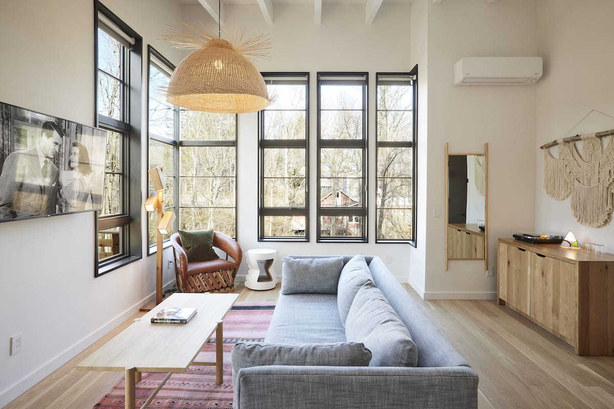 A modern living room with a grey sofa, a wooden coffee table, a wicker pendant light, and large windows with a view of trees.