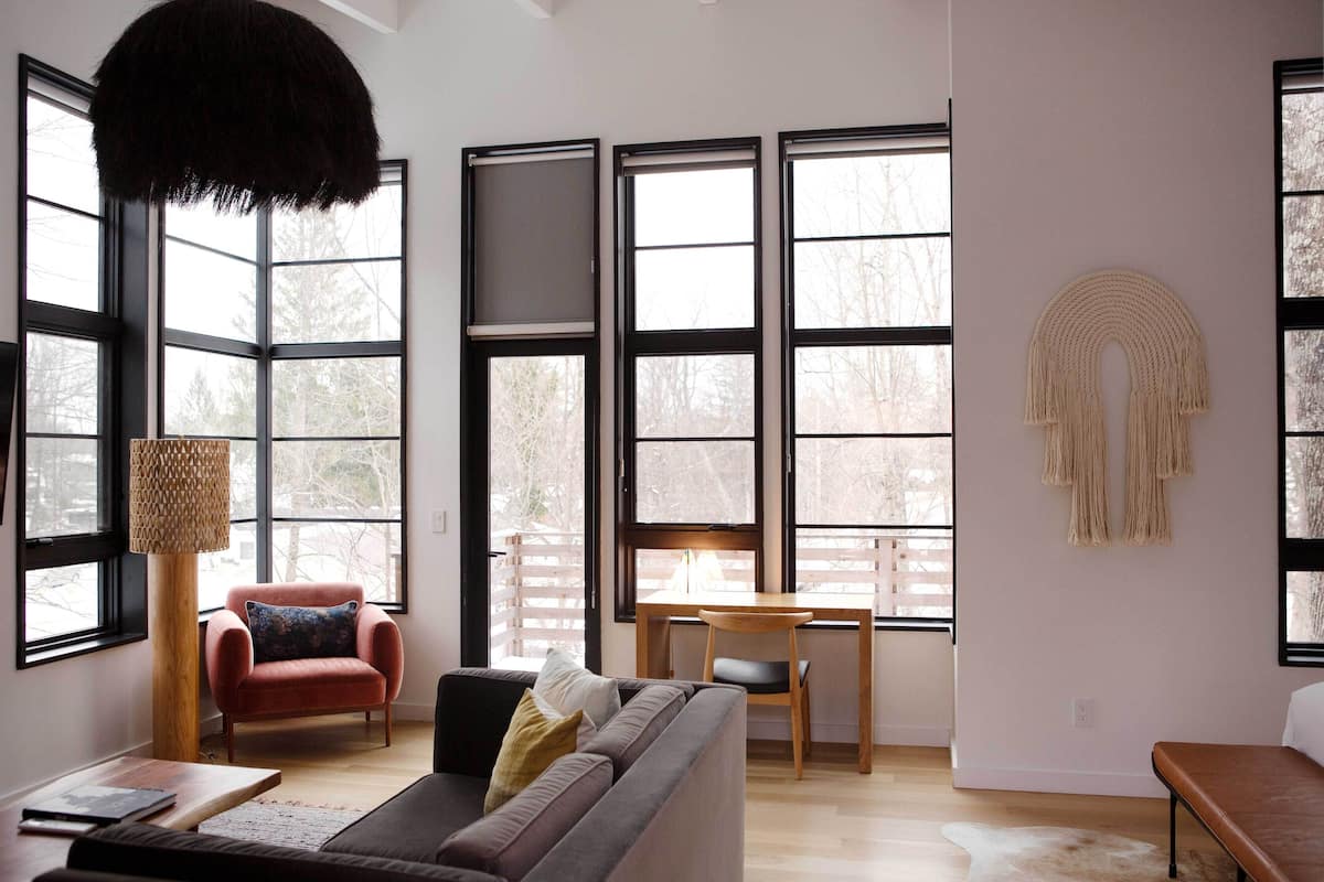 A modern living room with a sofa, armchair, and dining table. Large windows provide natural light and views of trees.