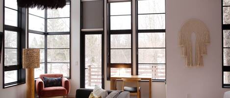 A modern living room with a sofa, armchair, and dining table. Large windows provide natural light and views of trees.