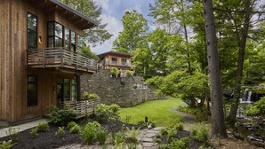 A two-story wooden house with a stone retaining wall, a stone pathway, and a small garden with various plants.
