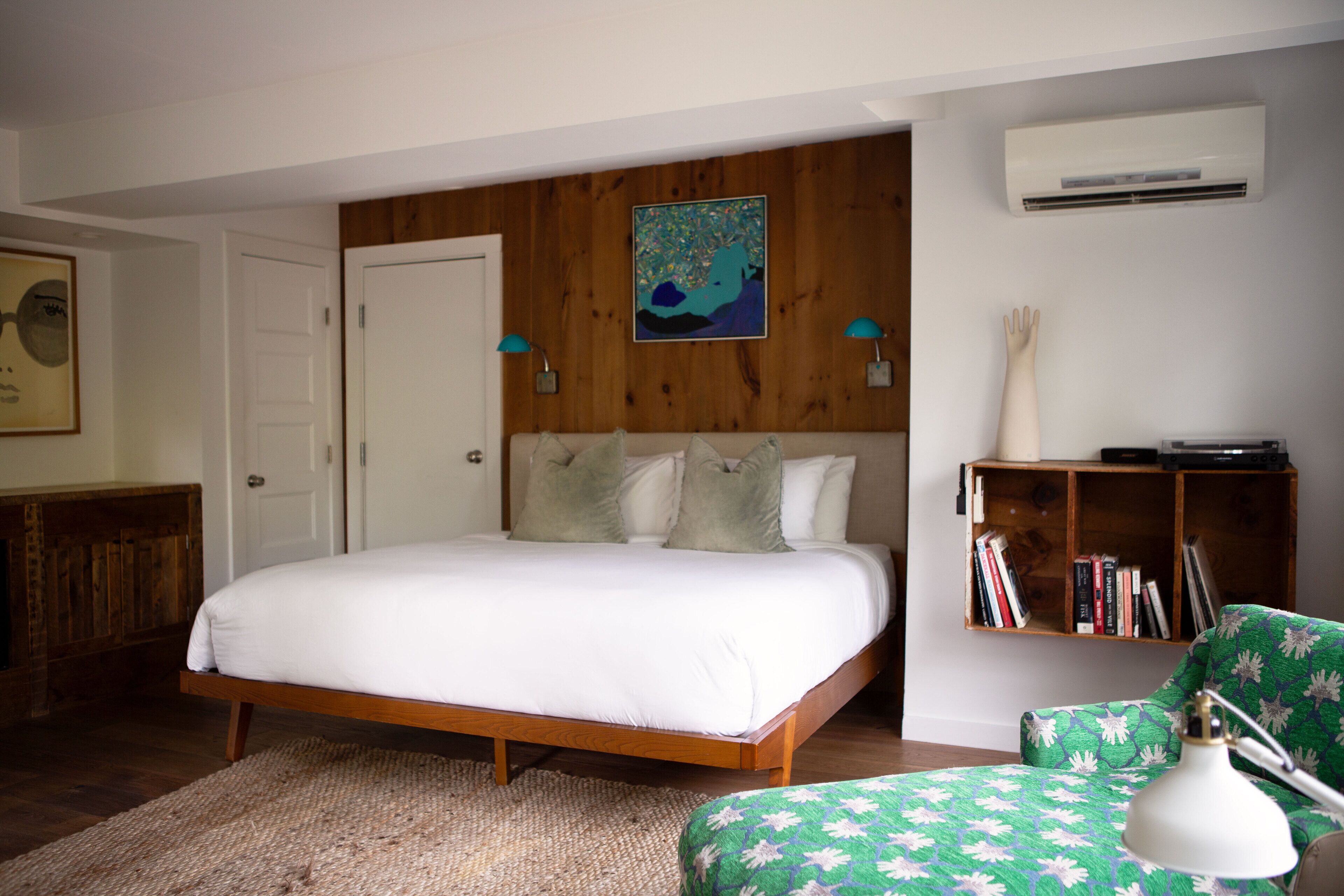 A bedroom with a wooden bed frame, a green patterned chair, a bookshelf, and an air conditioning unit.