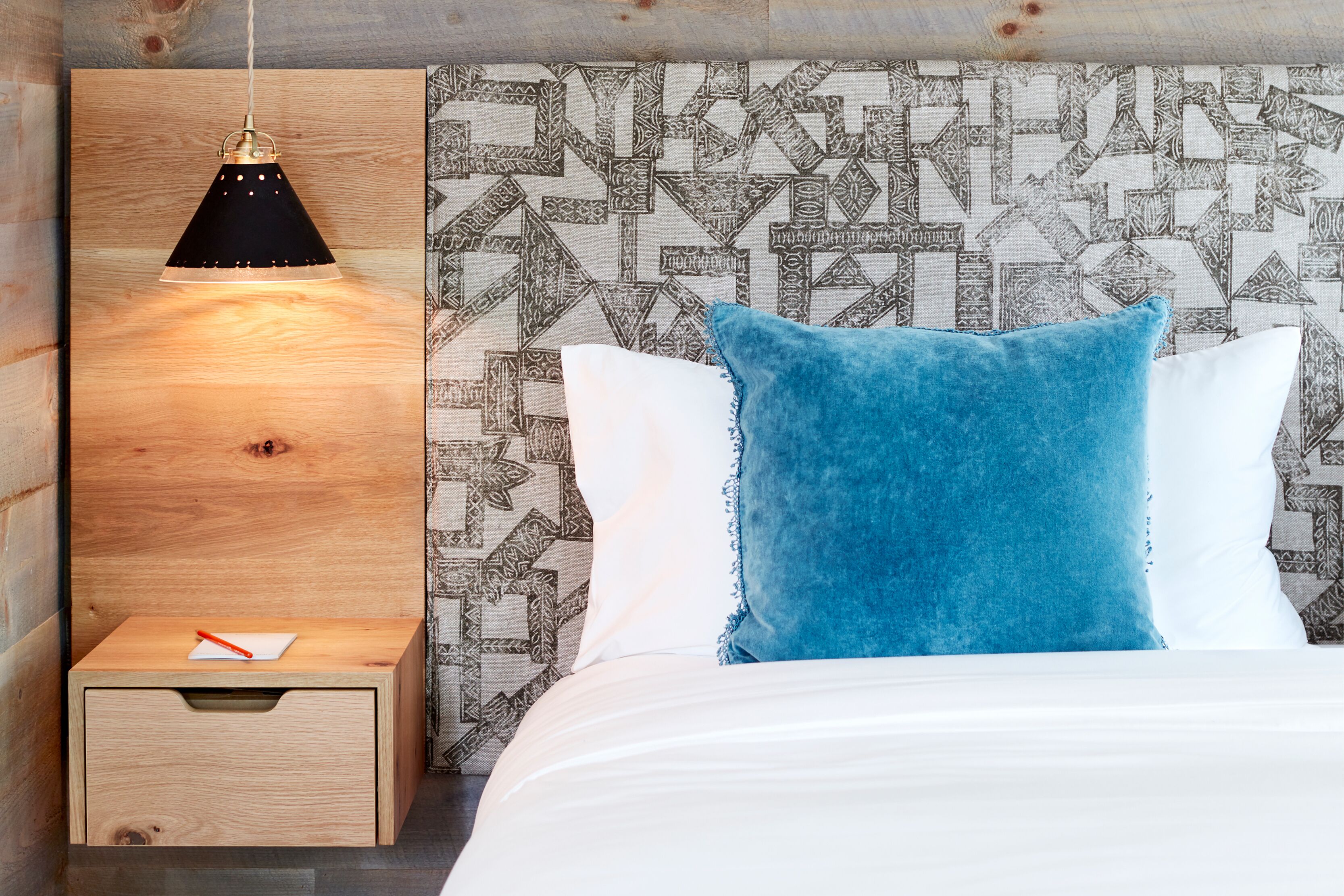 A bed with a blue pillow, a wooden bedside table with a pen, and a hanging lamp.