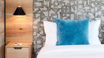A bed with a blue pillow, a wooden bedside table with a pen, and a hanging lamp.