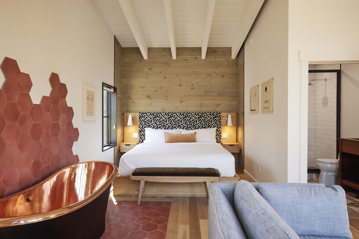 A modern bedroom with a large bed, a wooden headboard, a copper bathtub, and a blue sofa.