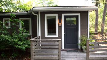 A small, single-story house with a dark front door, white-framed windows, and a wooden porch with steps leading to the entrance.