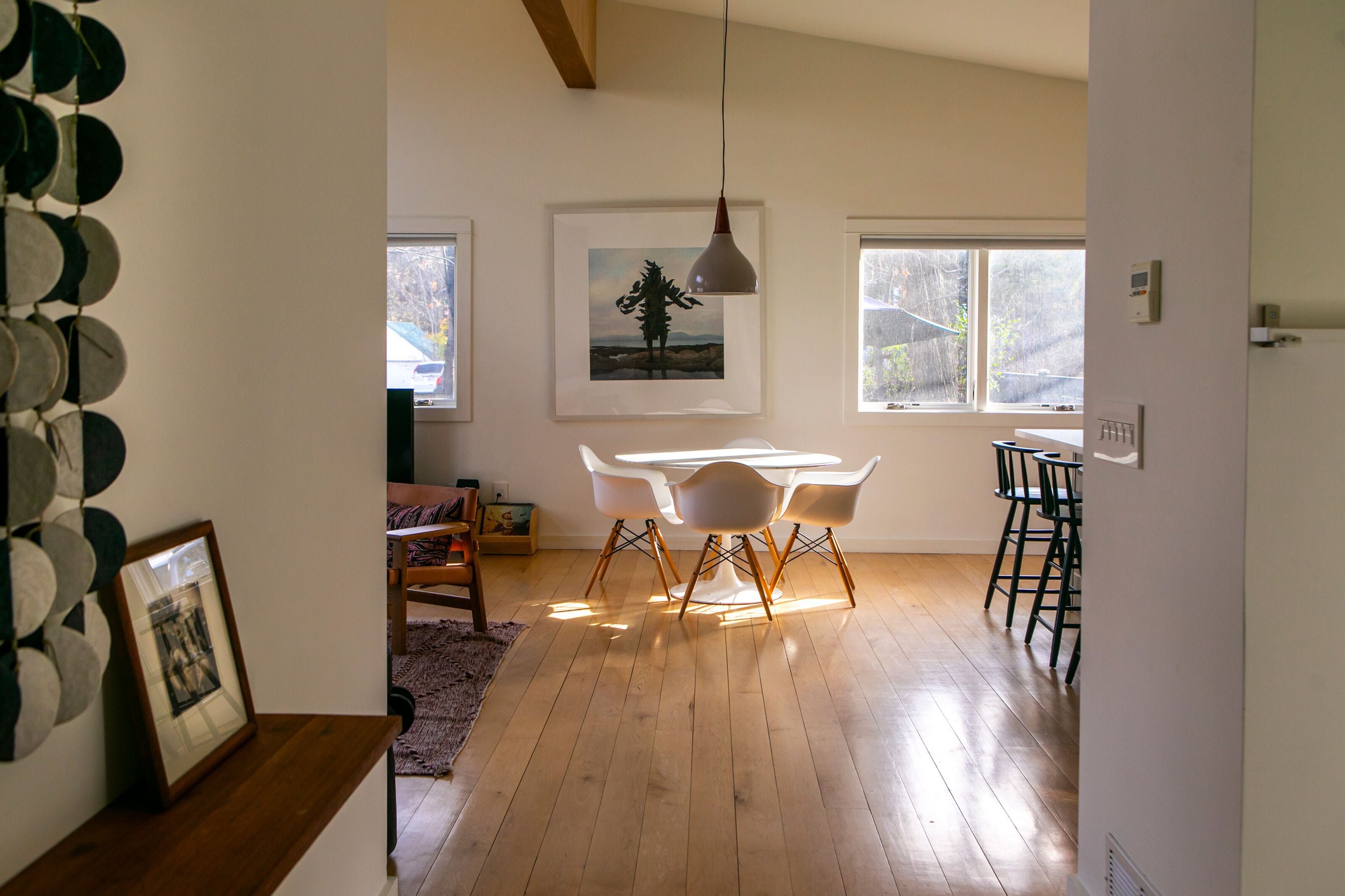 Una habitación amplia con suelo de madera, una mesa de comedor blanca con sillas, una lámpara colgante y un cuadro en la pared.