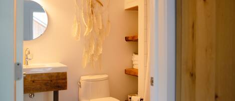 Un baño con inodoro blanco, tocador de madera, un atrapasueños y un espejo.