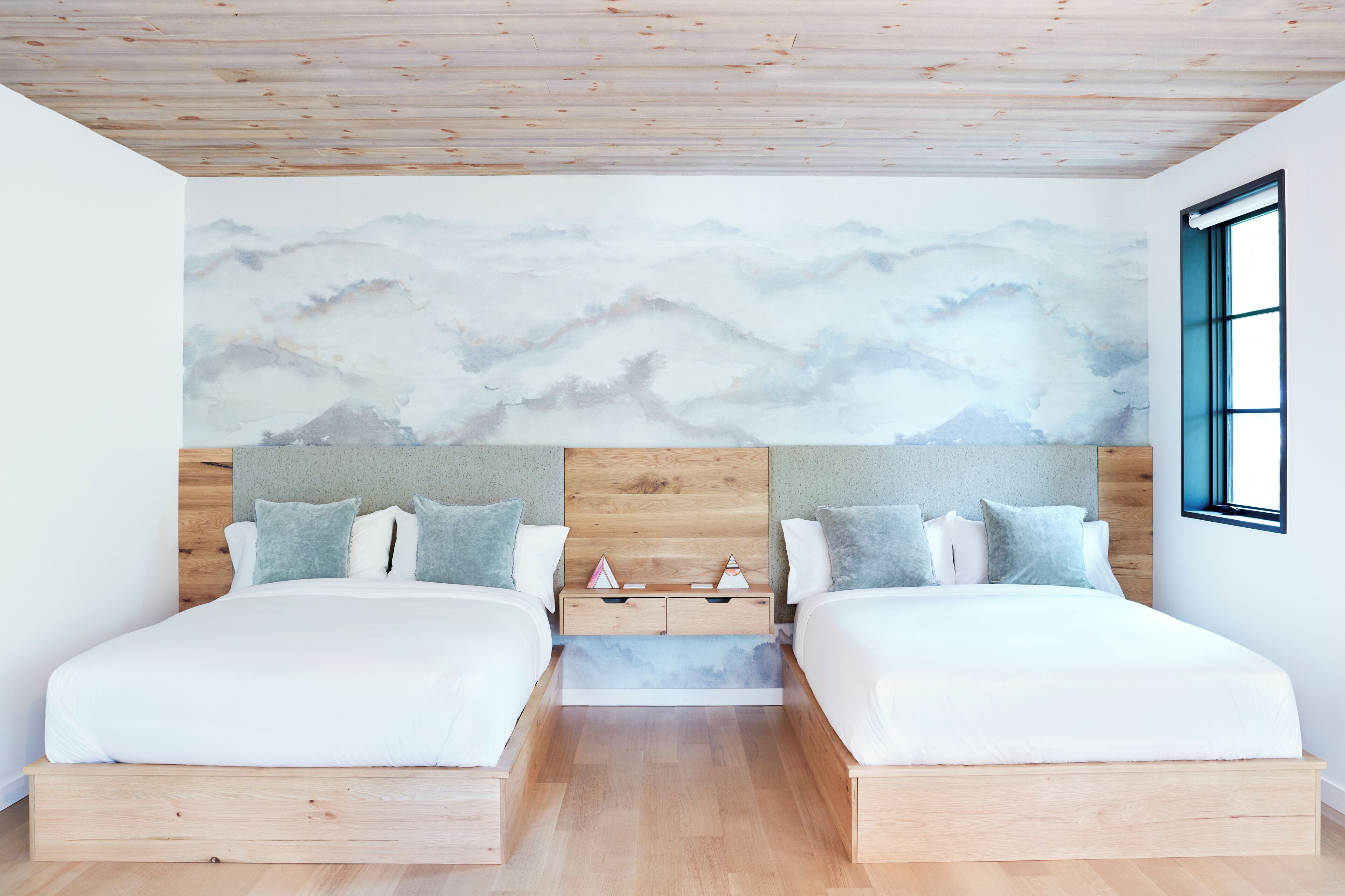 A modern bedroom with two beds, wooden headboards, a nightstand, and a large window.