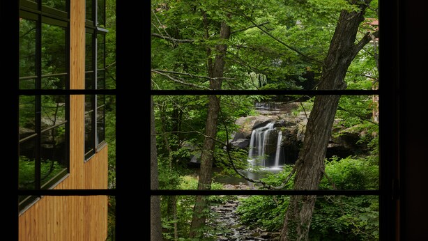 A wooden cabin with large windows overlooking a forested area with a waterfall.