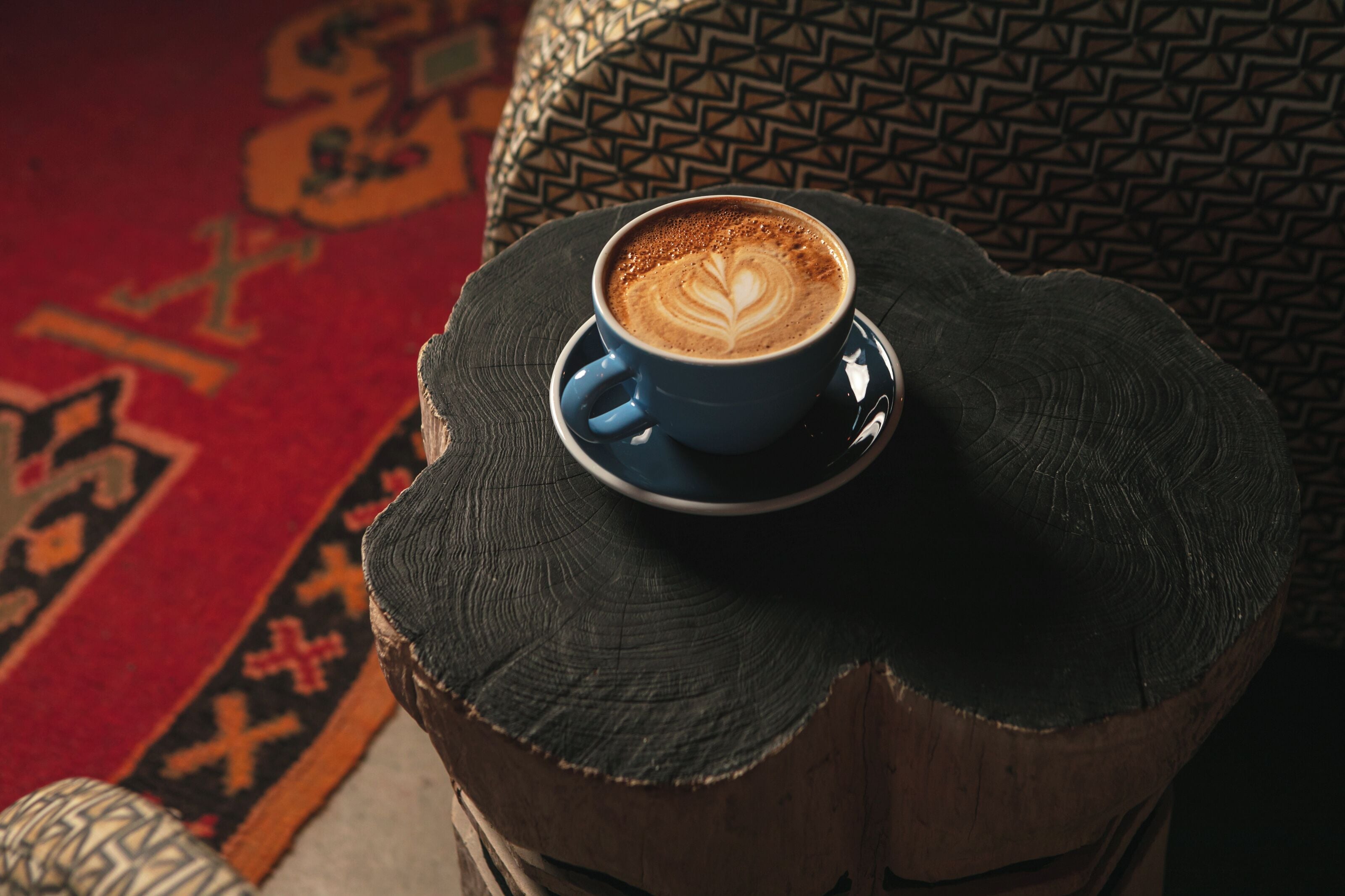 Eine Kaffeetasse auf einem hölzernen Baumstumpf, im Hintergrund ein Sitzkissen mit Muster.