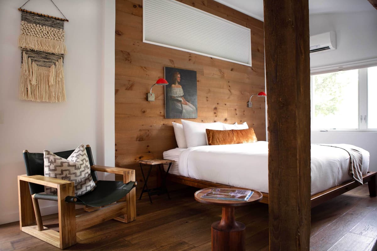 A wooden-framed room with a bed, a chair, a small table, and a wall-mounted macramé piece.