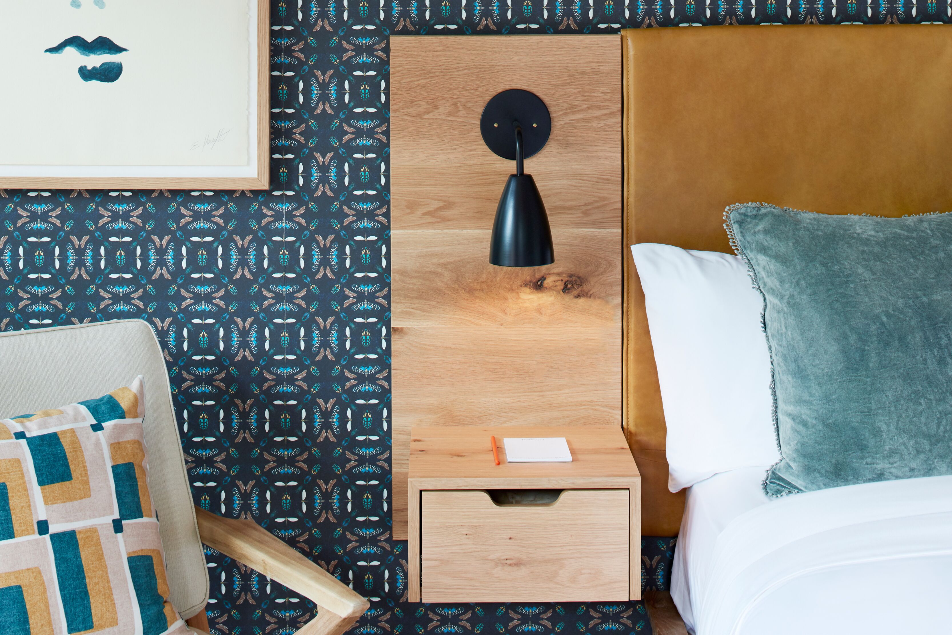 Habitación de hotel ordenada con una mesita de noche de madera, una mesilla con lámpara, una cama con almohada verde y una pared con diseño.