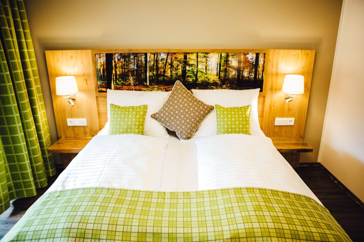 Habitación de hotel con una cama grande, dos mesitas de noche, cabecera con un mural de un bosque y dos lámparas de pared.