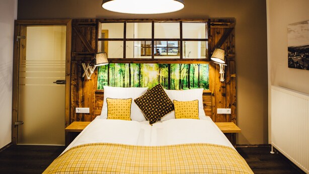 A bedroom with a large bed, wooden headboard, two bedside lamps, and a view of trees through a window. - Der Dorfwirt (Rechberg)