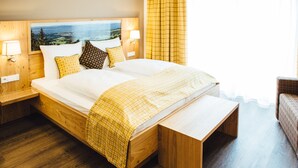 A neatly made bed with a view of the landscape, wooden headboard, two bedside lamps, and a bench at the foot of the bed. - Der Dorfwirt (Rechberg)