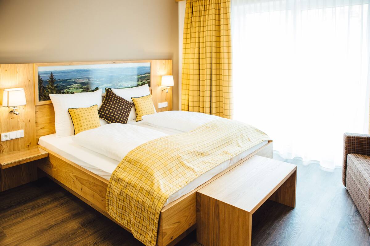 Una cama bien tendida con vistas al paisaje, cabecera de madera, dos lámparas de noche y un banco al pie de la cama.