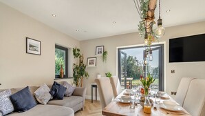 A modern living room with a dining area, featuring a sofa, a dining table set for four, and a large window offering a view of the outdoors.