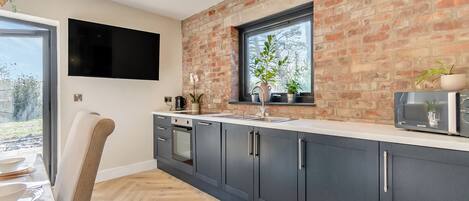 A modern kitchen with dark cabinets, a herringbone wood floor, a brick wall, a window, and a flat-screen TV mounted above the counter.