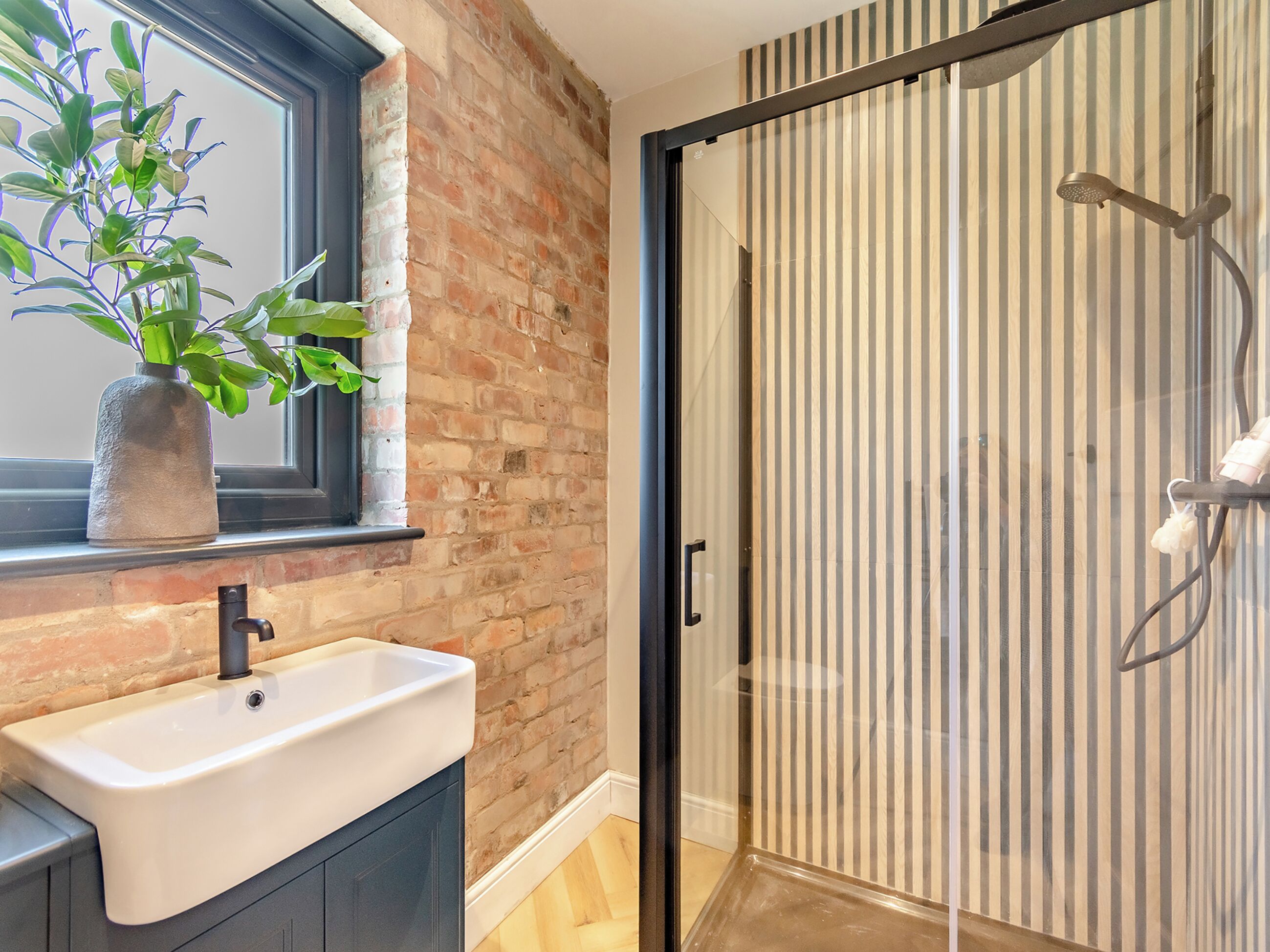 A bathroom with a white sink, a potted plant, a glass shower enclosure, and a brick wall.