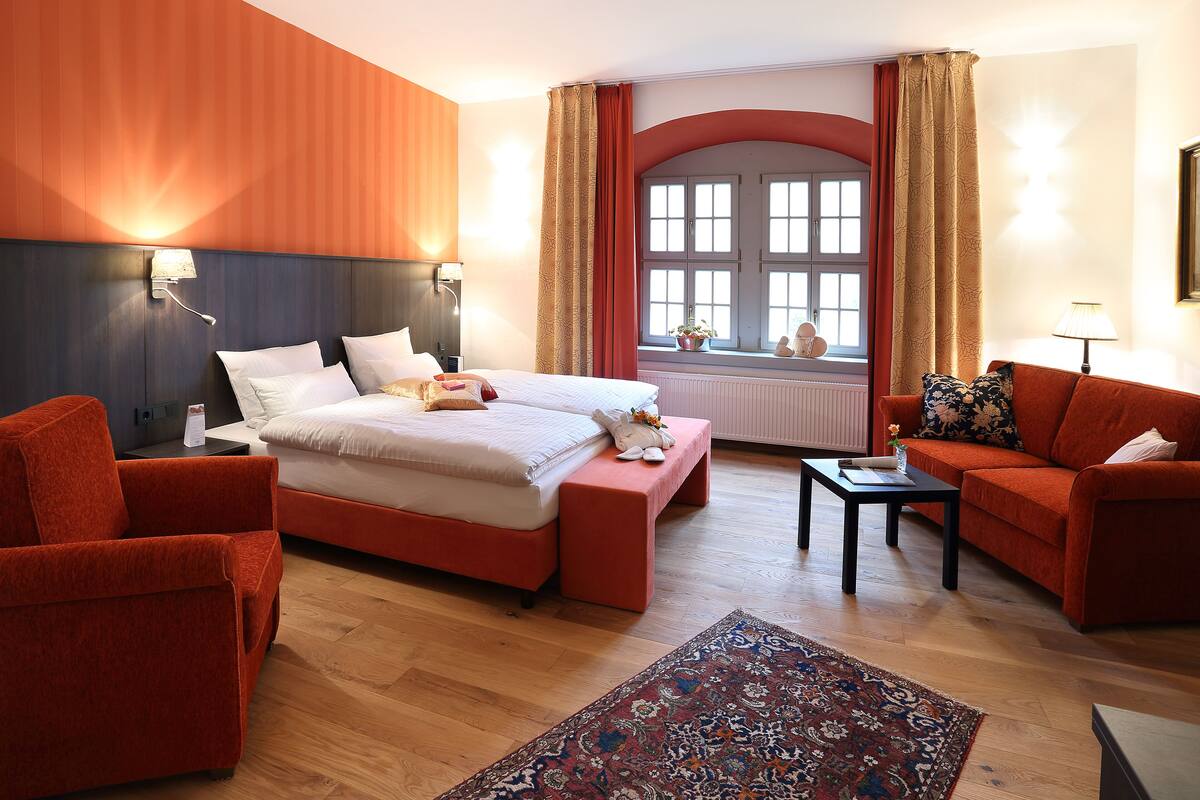 A hotel room with a large bed, two red armchairs, a small table, and a window with curtains.