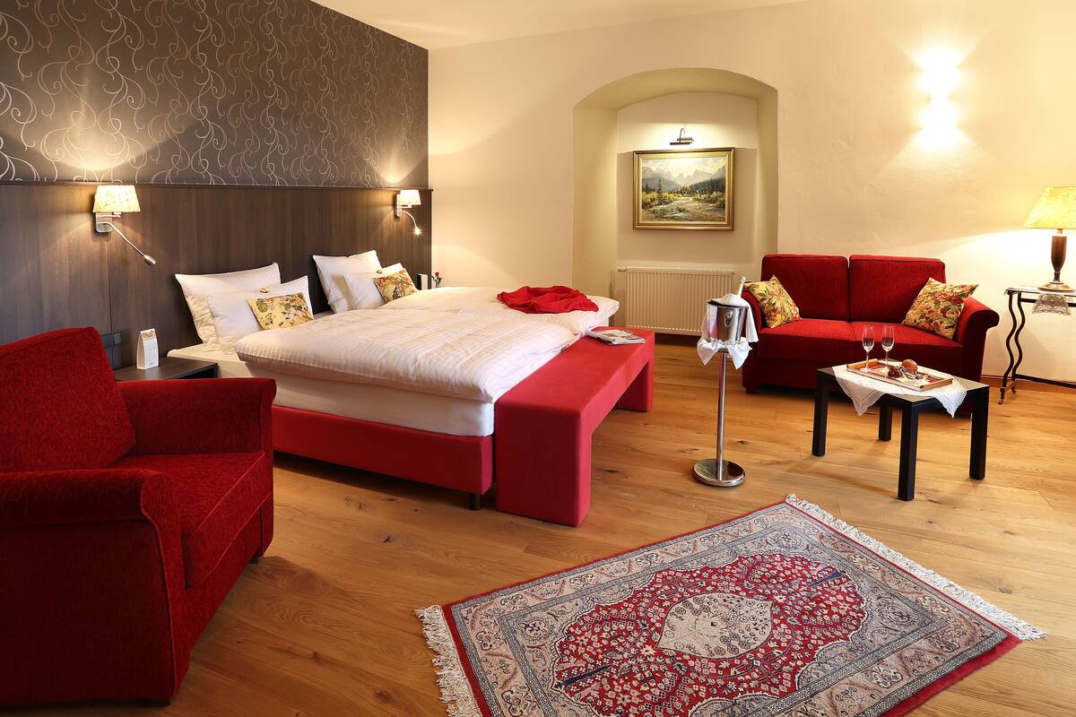 A hotel room with a large bed, a red sofa, a red armchair, a small table with glasses, and a patterned rug.
