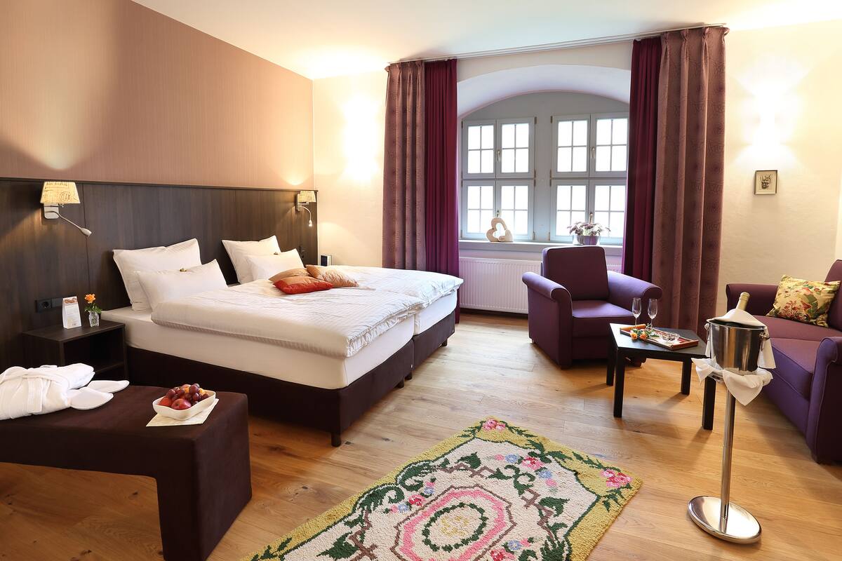 A hotel room with a large bed, a sitting area with a chair and a table, a small table with a pitcher, and a view of the outside through a window.