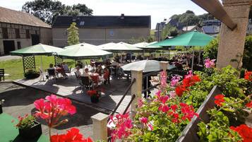 Ein Terrassenbereich im Freien mit mehreren Tischen und Stühlen unter grünen Sonnenschirmen, umgeben von Blumentöpfen und Blumen.