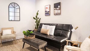 Ein Wohnzimmer mit einer schwarzen Ledersofa, einem Holzstuhl, einem kleinen Tisch mit einer Pflanze und einem großen Fenster.