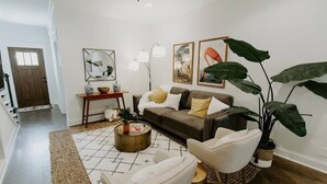 Un salon avec un canapé, des fauteuils, une table basse et une grande plante. Des tableaux encadrés ornent le mur, et une lampe est posée sur une table d’appoint.